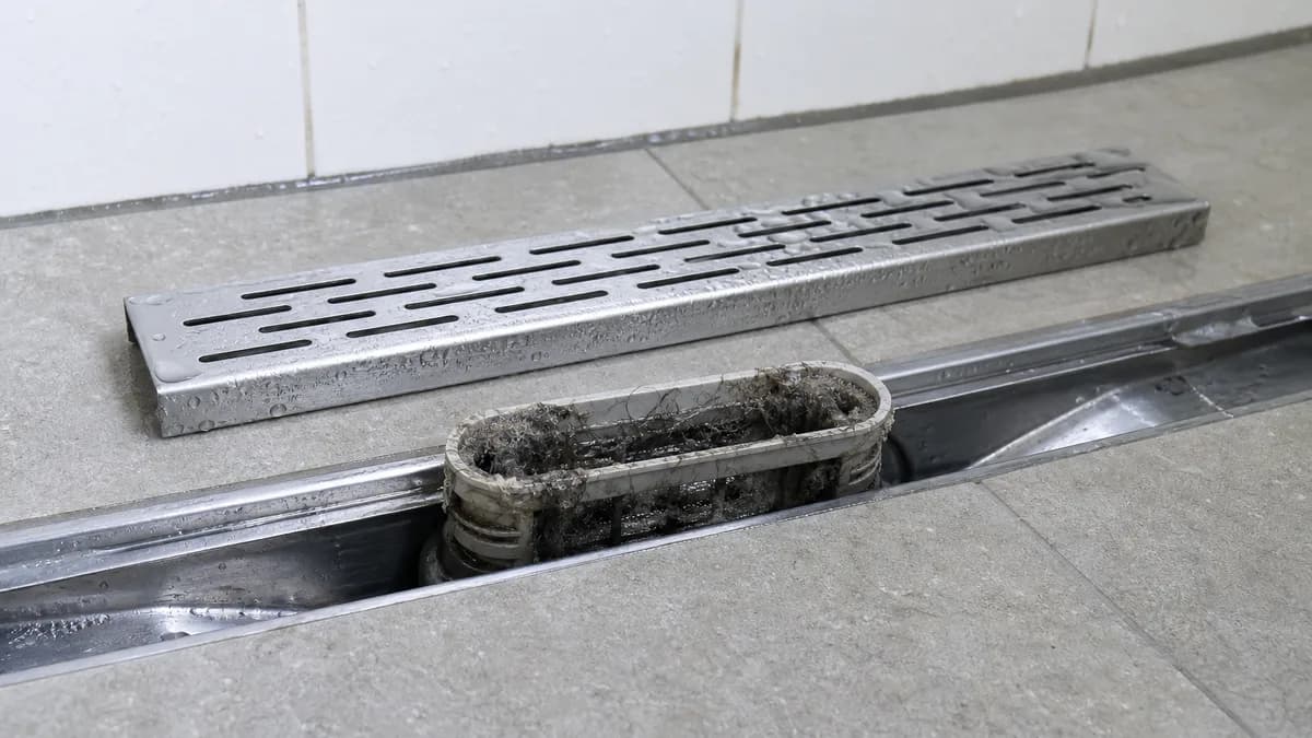 Abgenommener Edelstahl-Rost einer ebenerdigen Duschrinne, darunter der herausnehmbare Siphon-Einsatz mit Haar- und Schmutzablagerungen
